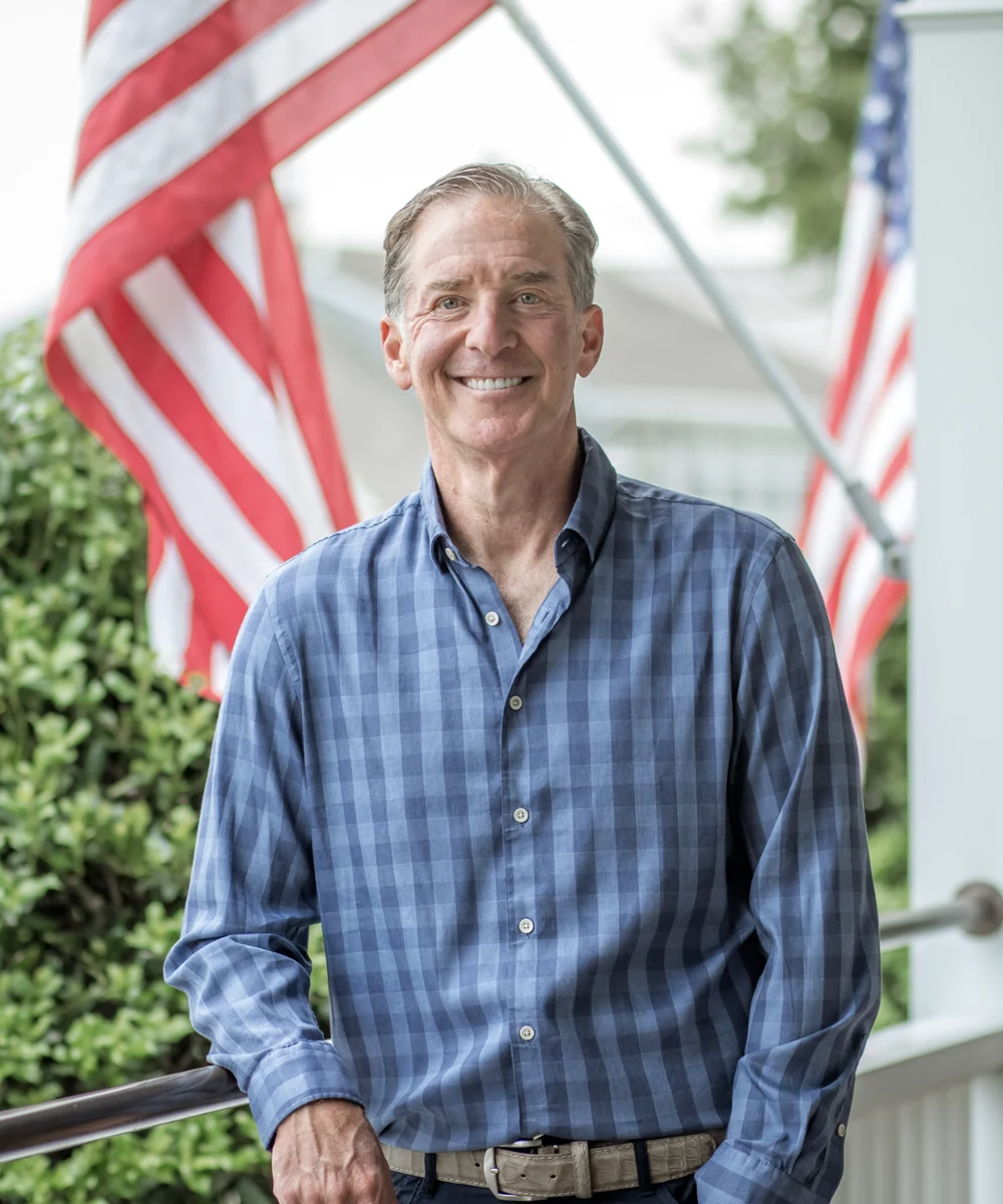 Mark Zides on a porch in a blue checked shirt, smiling, flags behind him.