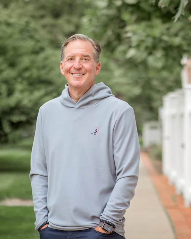 Mark Zides standing outdoors beside a white fence, in a grey hooded sweatshirt.